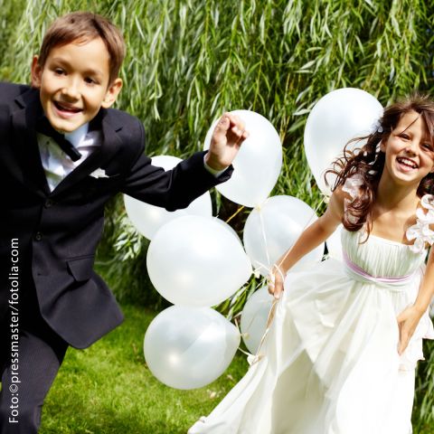 Kinder, Mädchen und Junge, festlich gekleidet laufen mit weißen Luftballons über eine Wiese