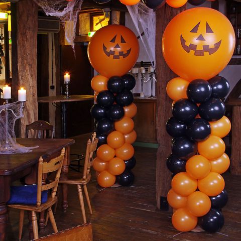 2 Luftballonsäulen in orange und schwarz, mit jeweils einem orangen Riesenballon als Abschluss, mit aufgedrucktem Kürbisgesicht.